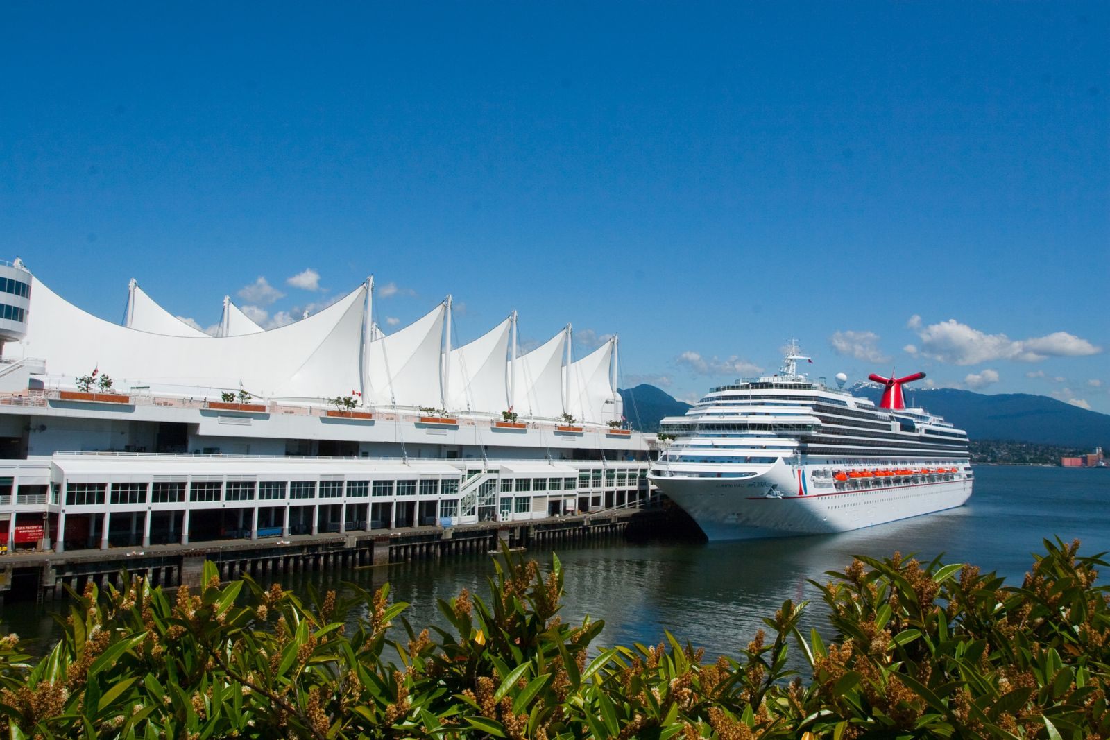 Cruise ship docked beside a white-sailed cruise terminal under blue sky